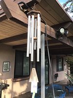 Full view of the wind chime hanging from a metal pole on a porch, showing all six metal tubes, wooden top frame, and windcatcher.