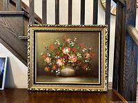 Full view of the blooming flowers oil painting in its ornate gold frame, placed on a wooden surface near a staircase.