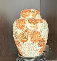 Front view of the vintage Japanese chrysanthemum ceramic jar with lid on a black carved stand (stand not included). Floral patterns in orange on an off-white background visible.