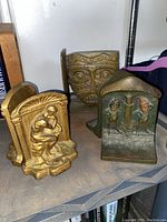 Photo showing three metal bookends with detailed designs: a gold one with a seated figure holding a child, a bronze-tone with two historical characters under an arch labeled 'The Pioneer' and 'The Farmer,' and another with a tribal face design.