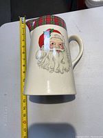 Ceramic pitcher with Santa Claus face and plaid ribbon handle, measuring approximately 7.5 inches tall.