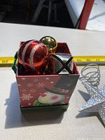 Box containing assorted Christmas ball ornaments and jingle bells with snowman design on box.