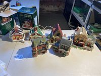 Six ceramic lighted Christmas village houses on a table, some with visible damage and cords for lights, arranged for display.