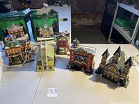Five porcelain lighted Christmas village buildings arranged on a table, some with boxes in the background. Includes a Fire Station, churches, and other village buildings.