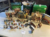 Full view of the porcelain Christmas village set including buildings and numerous figurines arranged on a table with original boxes in the background.