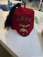 Photo of red Aleppo Shriners fez hat with black tassel and gold embroidery on a white surface.