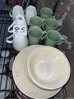 Photo showing 5 green mugs arranged in front of 4 white mugs with tags visible, along with bowls stacked nearby