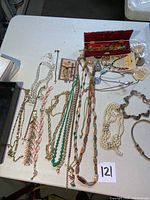 Photo shows an assortment of necklaces and pendants laid flat on a table, displaying a variety of styles including faux pearls, chains, and stone-like beads in multiple colors. Some pieces look vintage and worn.