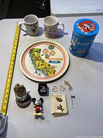 Photo showing all items on a white surface: two Disney mugs, California Disneyland souvenir tray, blue Looney Tunes tin, small clock on white base, Mickey and Minnie Mouse figurines, and multiple pieces of gold-tone Disney jewelry.