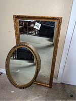 Photo showing large rectangular gold framed mirror with intricate floral and leaf patterns on frame, leaning against wall beside oval mirror.