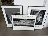 Three framed black and white Ansel Adams prints leaning against a wall: The American Wilderness in center, waterfall scene right, Half Dome moon scene left.
