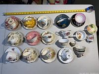 Full overhead view of 31 assorted tea cups and saucers laid out in rows on a white surface with measuring tape showing approximate dimensions.