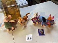 Winnie-The-Pooh plush toys Piglet, Tigger, Eeyore, Roo, Rabbit, and Pooh brooch set spread out on table.