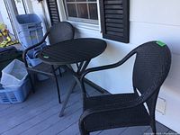 Two dark brown resin wicker-style patio chairs and a round black folding metal table on a covered porch.