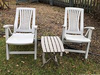 Two white plastic outdoor chairs with armrests and a small white folding table arranged on grass outdoors