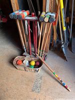 Wide photo showing the croquet set with mallets standing in a wooden rack, colored croquet balls in a wooden basket on the floor, and stakes included in the rack, all placed on wooden shed floor with other tools in background.