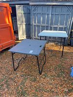 Photo showing two folding tables outdoors on a ground covered in brown leaves. Larger black table in foreground, smaller white table behind it.