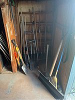 Photo showing an angled view of a collection of weathered garden tools leaning against a shed wall, including shovels, rakes, and a broom.