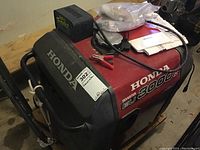 Overall view of red and black Honda EU3000is inverter generator on wooden pallet with Battery Tender Plus and manuals on top