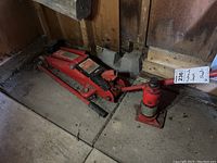 Two red hydraulic jacks on floor of shed, one a Motomaster trolley jack the other a bottle jack.