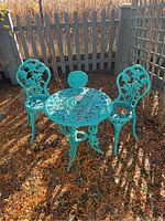 Full view of turquoise bistro set with one round table and two chairs outdoors on brown leaves, next to gray fence.