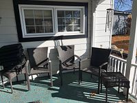 Three dark brown resin wicker patio chairs with cushions arranged on a porch next to a window, alongside two matching stools stacked to the side.