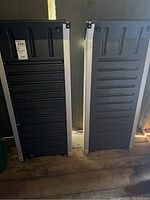 Two black plastic pet ramps with silver side rails standing vertically inside a shed.
