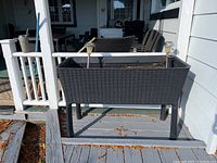 Front view of the black resin planter box on a porch, showing the rectangular shape and woven texture.