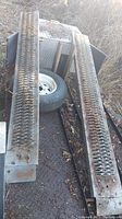Pair of metal vehicle ramps with grated tread surfaces lying on a truck bed outdoors with visible rust.