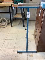 Full view of metal folding roller stand with blue frame and rectangular roller top. Visible adjustment knob on the vertical column.