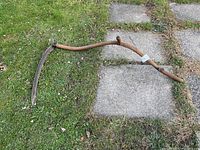 Full view of the vintage scythe lying on grass and concrete stepping stones, showing the bent long wooden handle and attached curved metal blade.