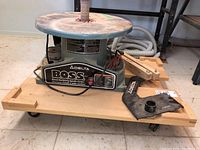 Front view of Delta BOSS oscillating spindle sander mounted on wooden dolly, showing power cord, power switch, and accessories.