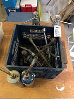 Photo showing multiple metal wrenches and two handheld blow torches with metal nozzles inside and beside a black plastic crate on a wooden surface.