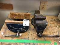Photo of two workshop vises on a workbench, one blue with wooden jaw covers and smaller, the other larger metallic grey vise.