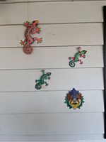 Photo of four colorful metal wall art pieces including three geckos and a lion face on white panel siding.