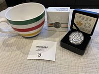 Photo showing large Hudson's Bay striped mug, unopened silver coin box, and coin in black clamshell case with certificate visible inside lid.