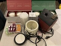Photo showing two boxed sets of holiday scented candles with three candles each (one missing), two ceramic Scentsy plug-in warmers—one cream with moose design and one red house-shaped warmer with star cutouts—additional scented wax refills, and a white textured pillar candle.