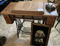 Singer wooden sewing table with open drawer, showing wear, scratches and missing drawer pull. Old iron and trivet on tabletop and framed dog acrylic on velvet leaning against table.