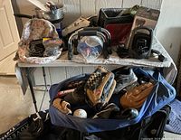 Wide view of table and bags showing multiple baseball gloves, bats, softballs, shoes, coolers, backpacks, and bags of sports and travel items.