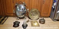 Photo of all six vintage items arranged on wooden floor: oval brass convex mirror, brass embossed tavern plate, house blessing plaque, Belgian copper simmer pot, holy water font, and electric wall sconce.