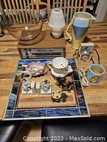 Photo showing vintage collectibles including a blown glass bowl, ceramic pitcher, vintage camera, shaving mug, and porcelain figurines on handmade stained glass frame.