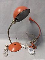 Side view of the orange desk lamp with dome shade showing flexible metal neck and base with power cord and switch, alongside the orange clamp lamp with small shade and clamp base.