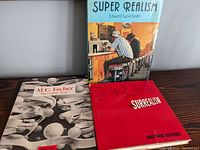 Three modern art books displayed on a dark wooden surface: 'Super Realism' showing a diner scene on the cover, 'Surrealism and Their Heritage' in red cover, and 'M.C. Escher The Graphic Work' with a monochrome surreal graphic design.