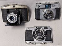 Three vintage cameras displayed on a table: folding Ansco camera, Ilford Sportsman, and Fujica 35mm camera.
