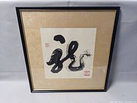 Framed Asian calligraphy print with black brush strokes and red seals inside beige mat and black frame.