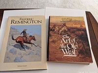 Two hardcover art books side by side on a surface. Left book titled 'Frederic Remington' with cowboy horse illustration on cover. Right book titled 'The Art of the Old West' with Western cattle drive cover art.