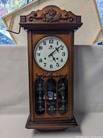 Full front view of a wooden wall clock with carved floral decoration above the clock face and glass panels below. Clock includes a winding key.