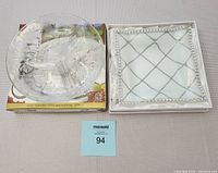 Photo of two brand new serving plates: clear glass divided plate with etched design on top of original box, and square white ceramic plate with silver decoration.