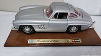 Side view of silver Mercedes 300 SL model car mounted on brown wooden base with plaques showing "Mercedes 300 SL (1954)" and Bburago logo.