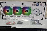 Front view of the Thermalright Frozen Prism 360 White ARGB cooler box showing the 3 ARGB fans and CPU block with logo and LEDs. Hardware and mounting plates are laid out in front.